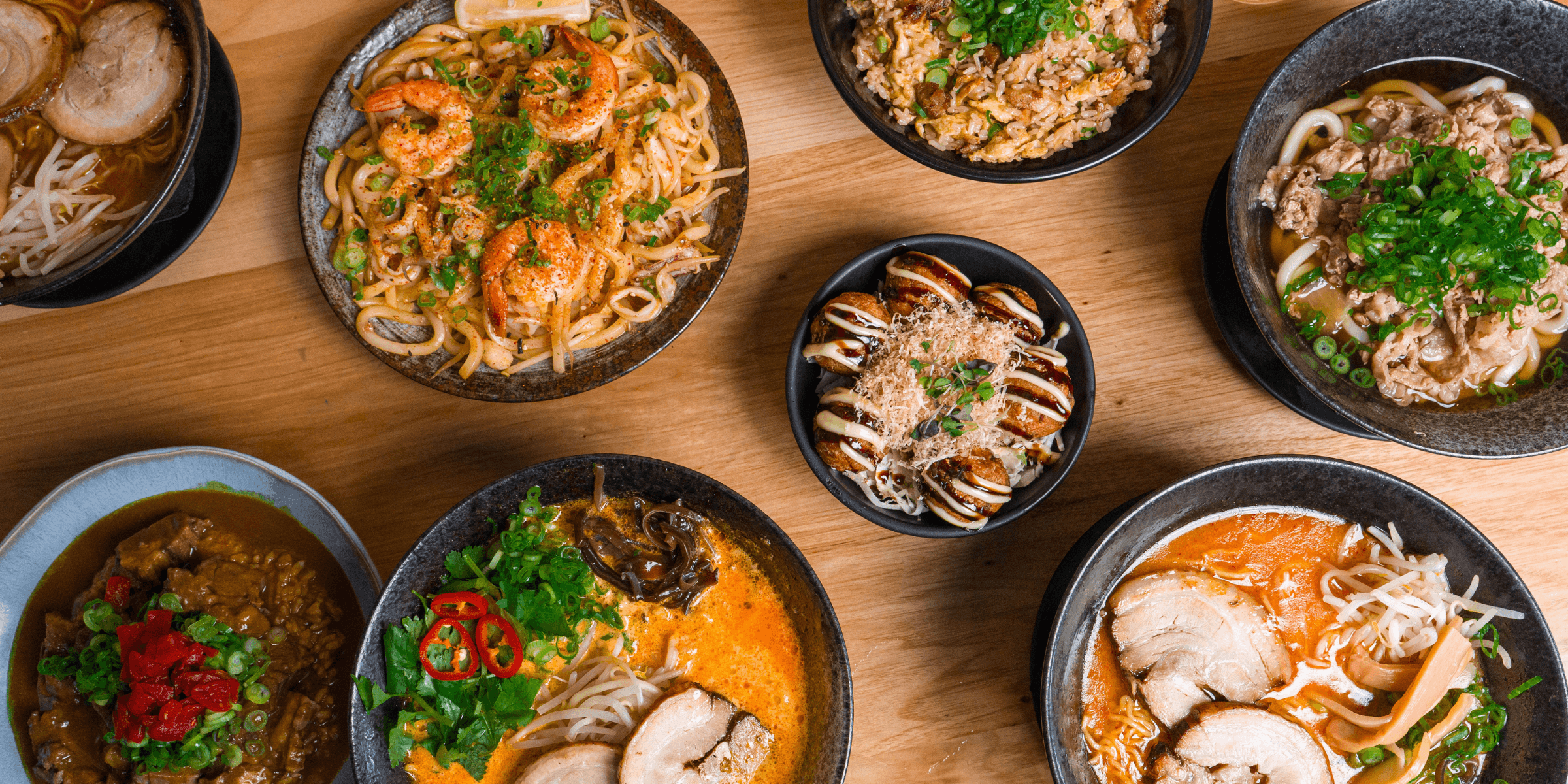 An overhead spread from Kin Craft Ramen & Izakaya featuring tom kha ramen, beef belly curry, takoyaki topped with sauce and bonito flakes, beef udon, shrimp stir fried udon, fried rice, and assorted ramen bowls arranged across a wooden table.