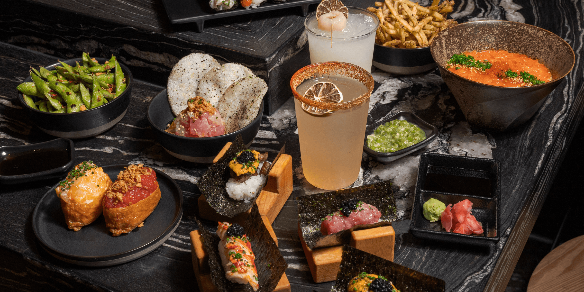 A spread from ROL Hand Roll Bar featuring assorted hand rolls topped with fresh fish and caviar, spicy edamame, appetizers, fries, dipping sauces, and signature cocktails arranged on a dark marble table.