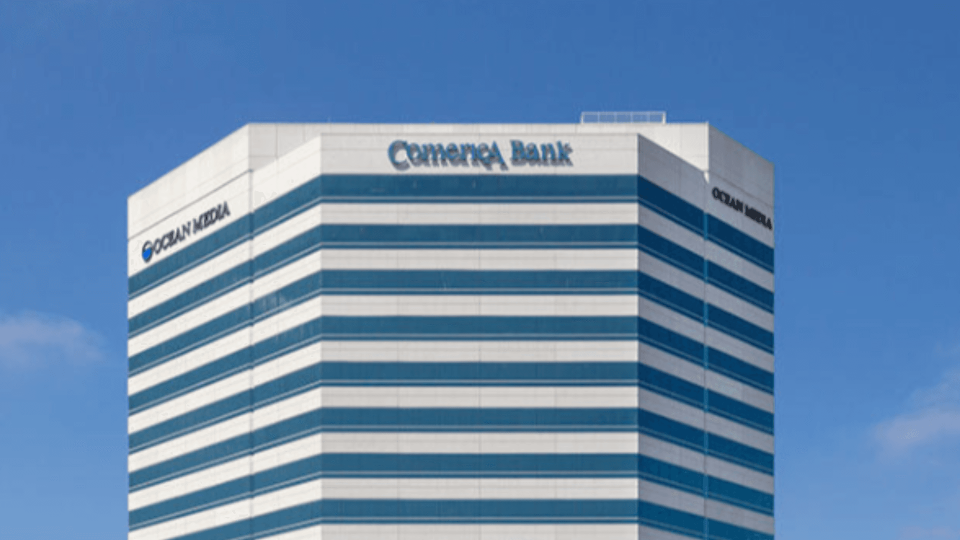Exterior of Kei Concepts headquarters building featuring a modern high-rise office tower with blue and white horizontal stripes against a clear blue sky.