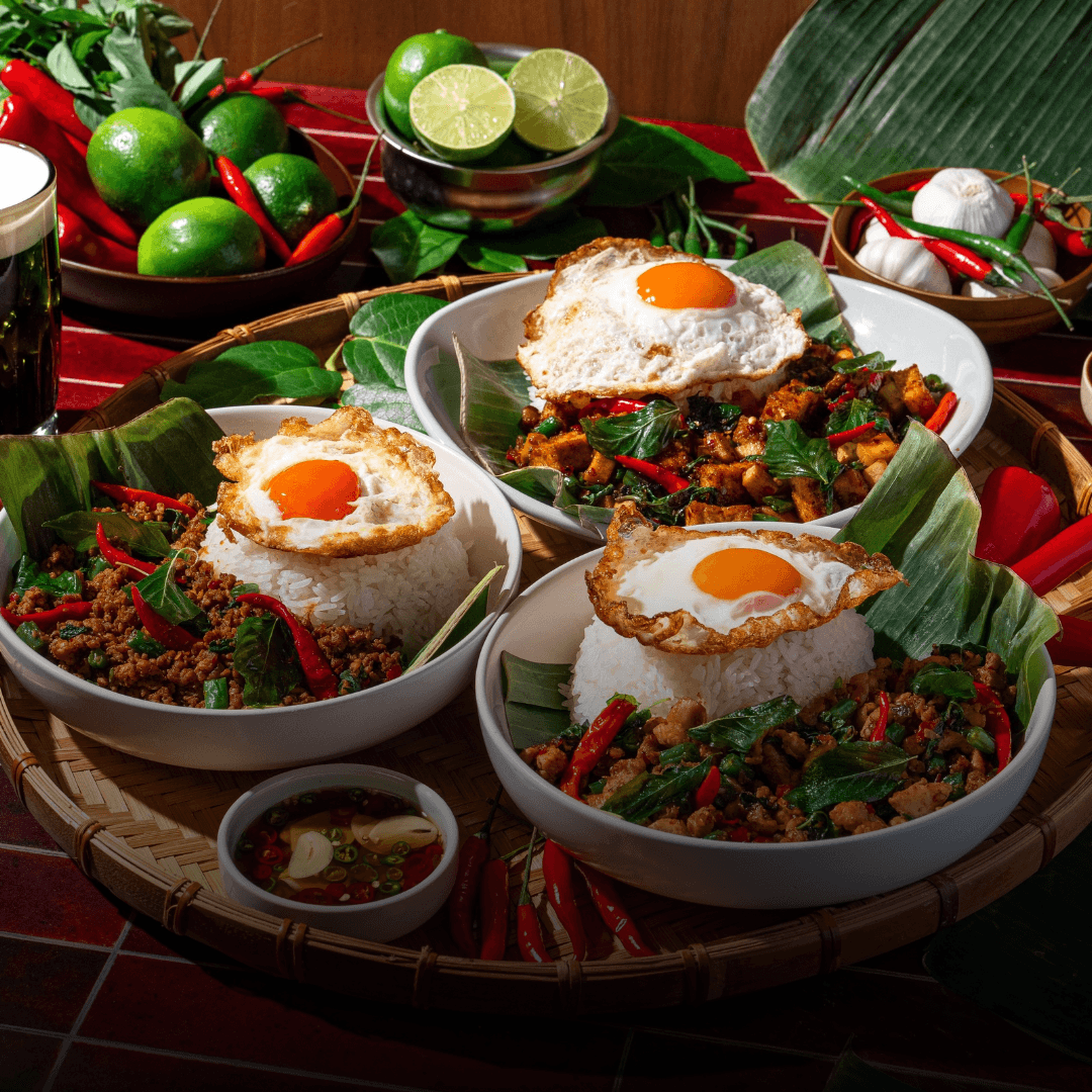 Holy Heat Pad Kra Pow bowls with spicy Thai basil stir-fry, white rice, and crispy fried eggs, surrounded by fresh chilies, limes, garlic, and Thai-inspired ingredients.