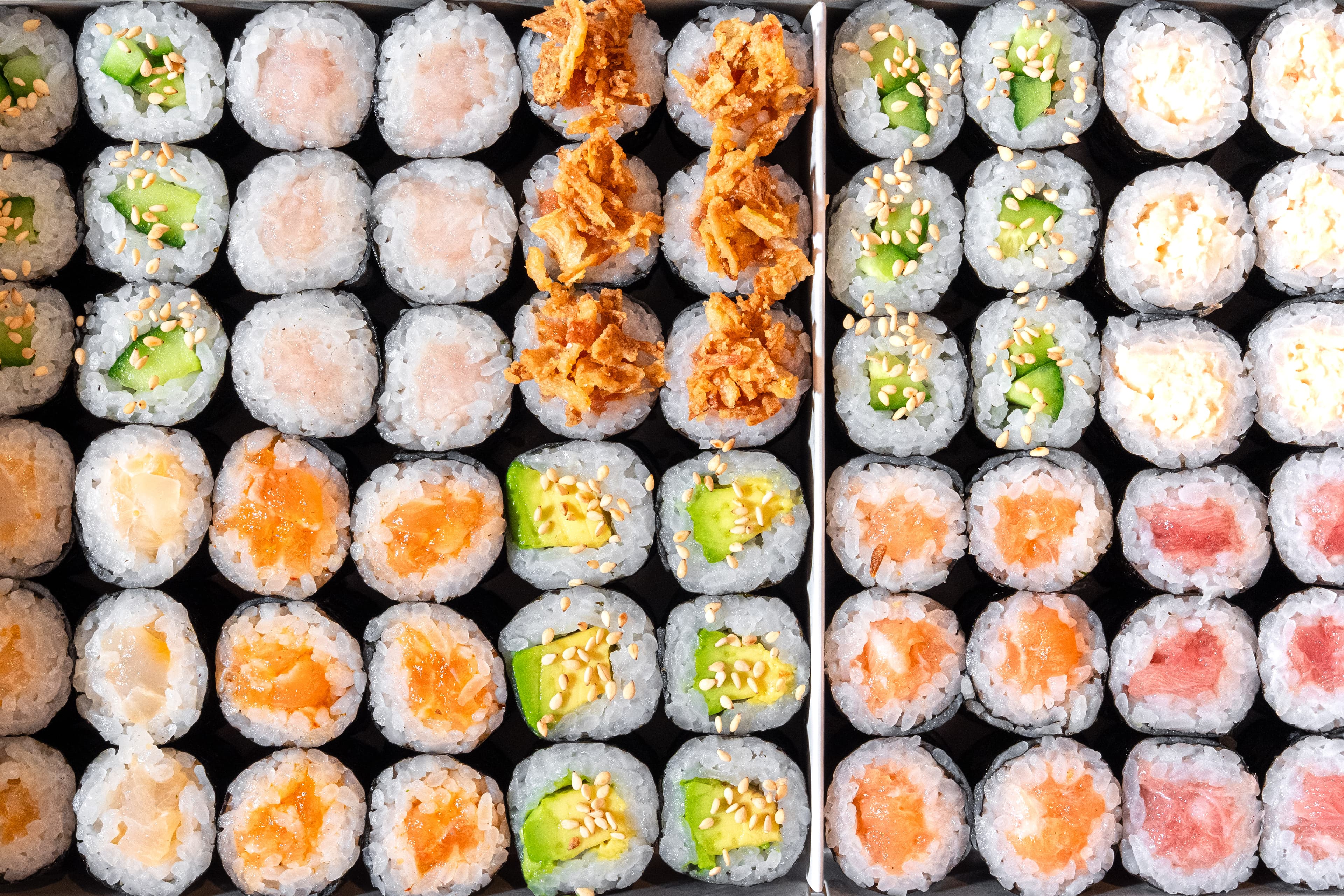 Overhead close-up of assorted sushi rolls arranged neatly in black trays, featuring avocado, cucumber, salmon, tuna, creamy fillings, sesame seeds, and crispy toppings