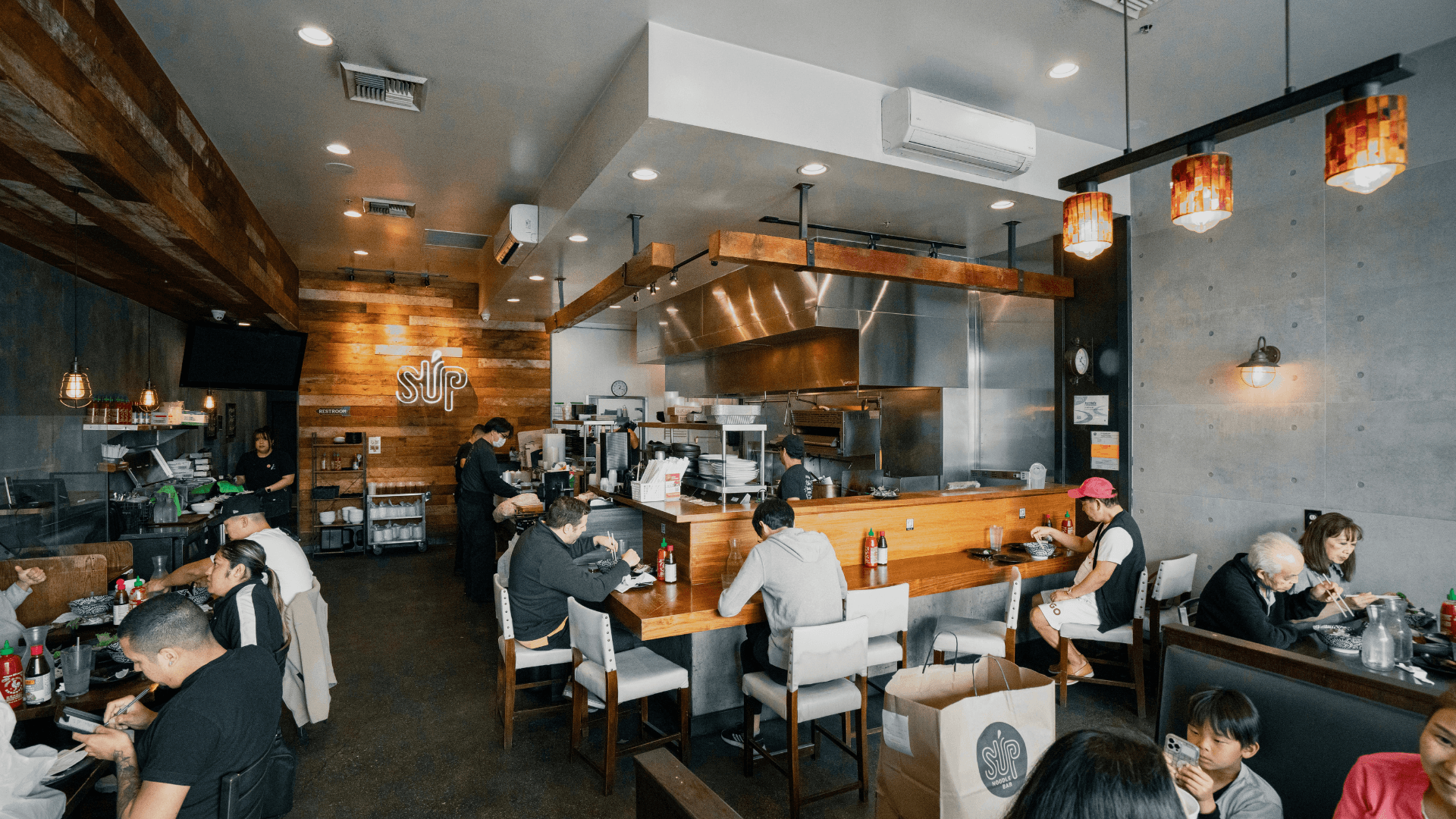Interior of SUP Noodle Bar’s Buena Park location featuring a warm modern dining space with wood accents, open kitchen seating, pendant lighting, and guests enjoying meals throughout the busy restaurant.