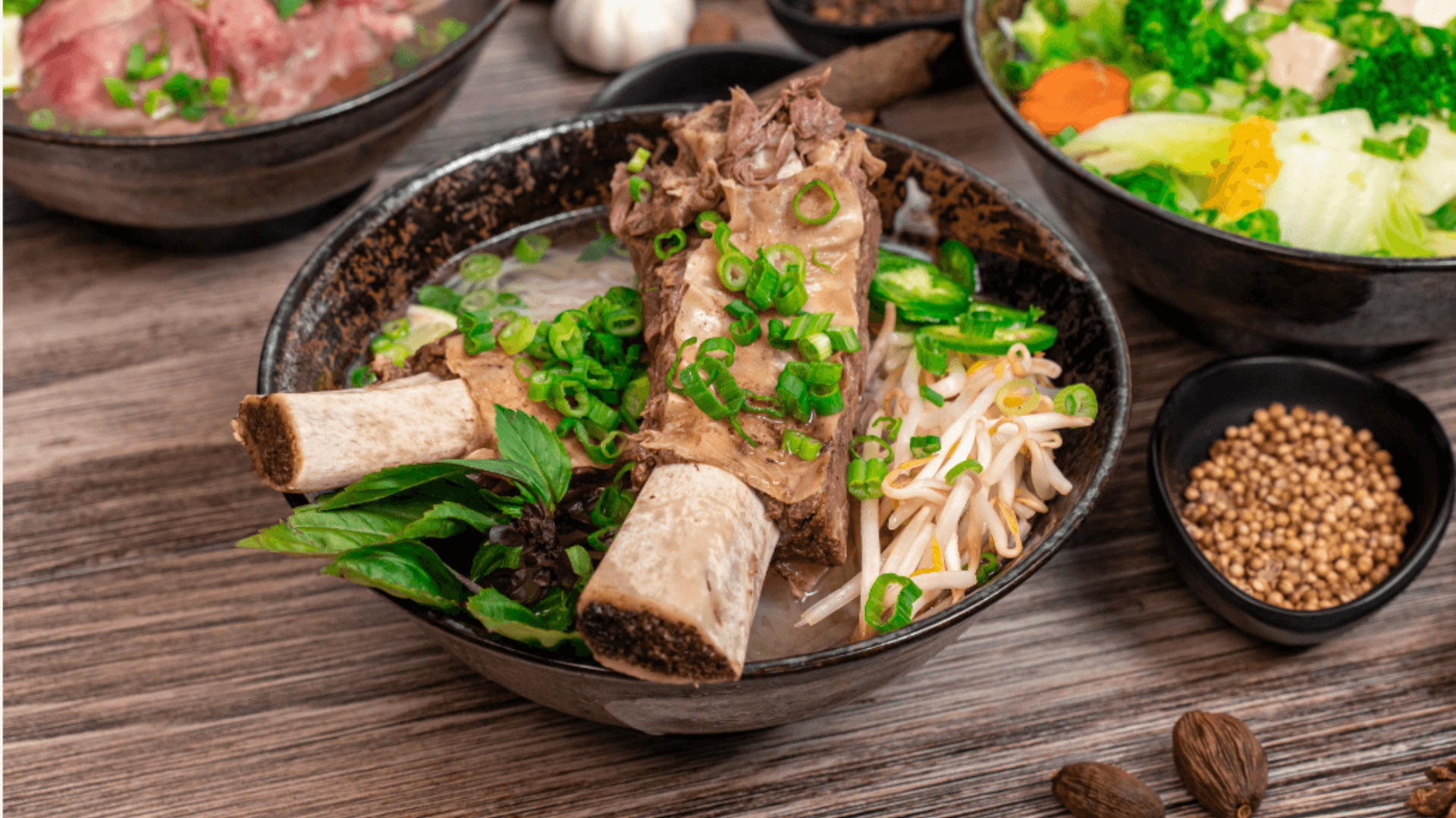 Close-up of SUP Noodle Bar’s rib bone pho featuring large beef rib bones in a rich broth with rice noodles, bean sprouts, scallions, fresh basil, and jalapeños served in a rustic black bowl.