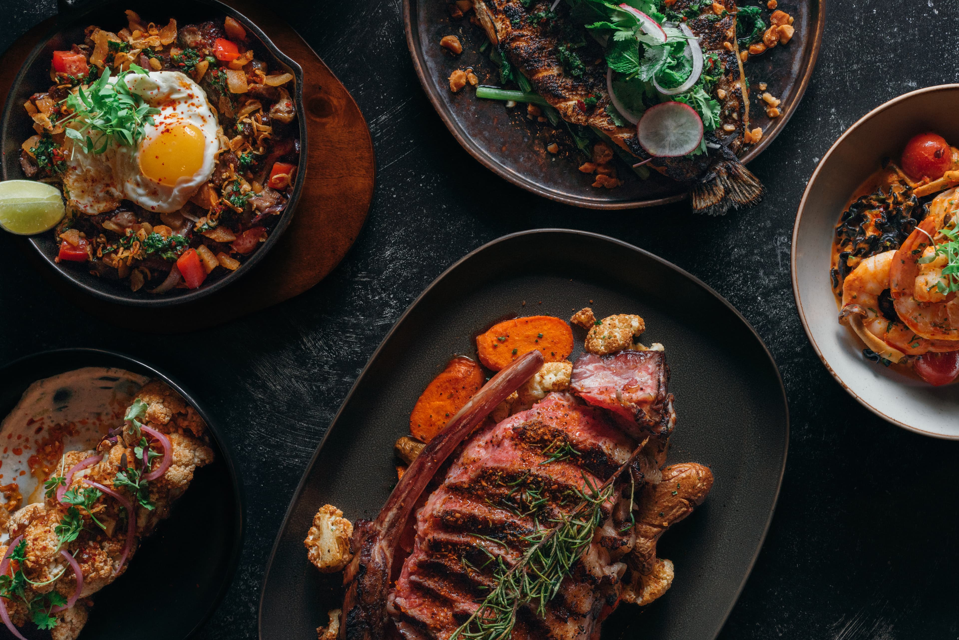 An overhead spread from GEM Dining featuring grilled tomahawk steak with roasted vegetables, fried rice topped with a sunny-side egg, whole grilled fish, shrimp pasta, and elevated Asian fusion dishes plated across a dark modern table setting.