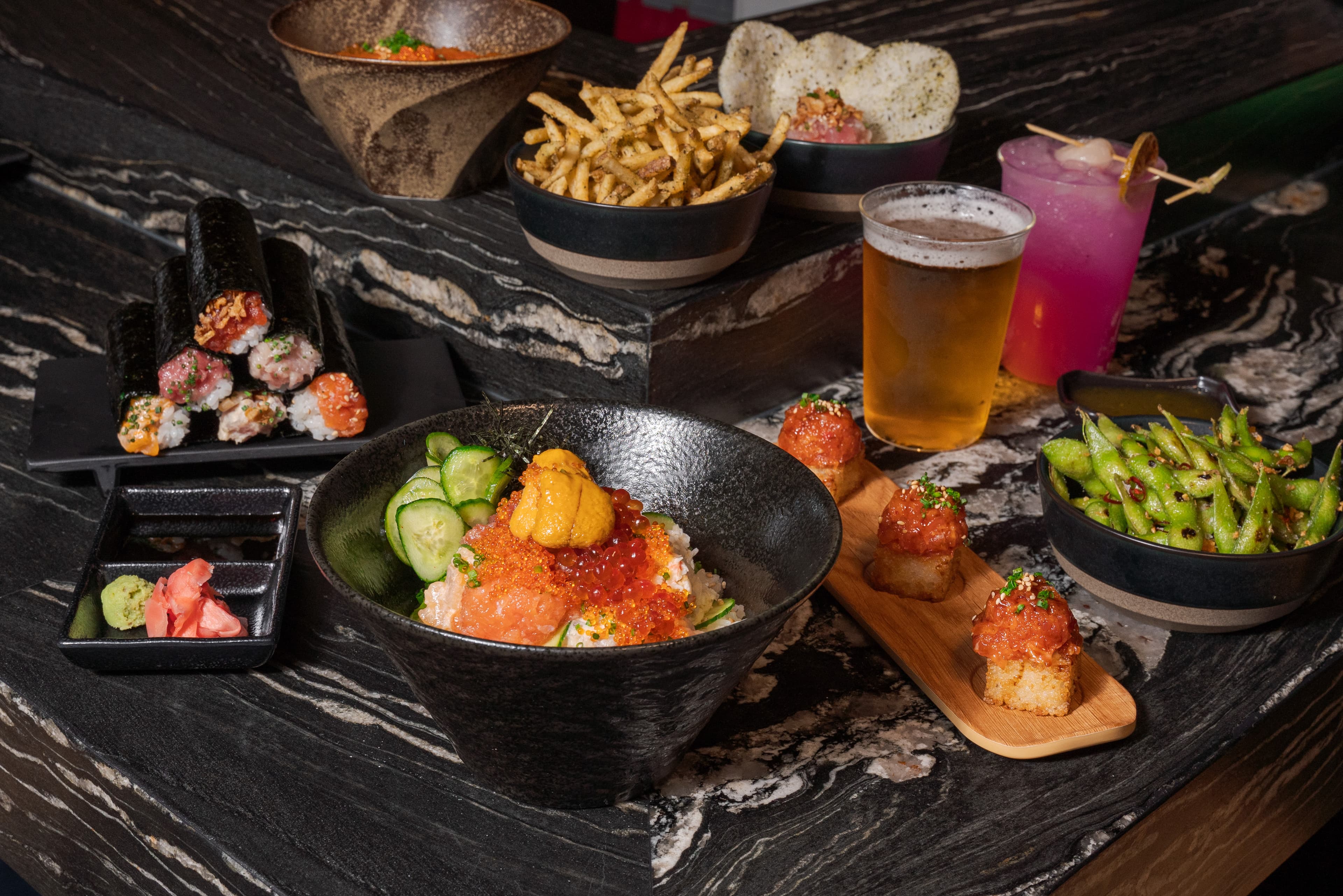 A spread from ROL Hand Roll Bar featuring kaisendon topped with salmon, ikura, and uni, assorted hand rolls, spicy edamame, spicy tuna crispy rice, seasoned fries, sushi appetizers, and drinks arranged on a dark marble table.