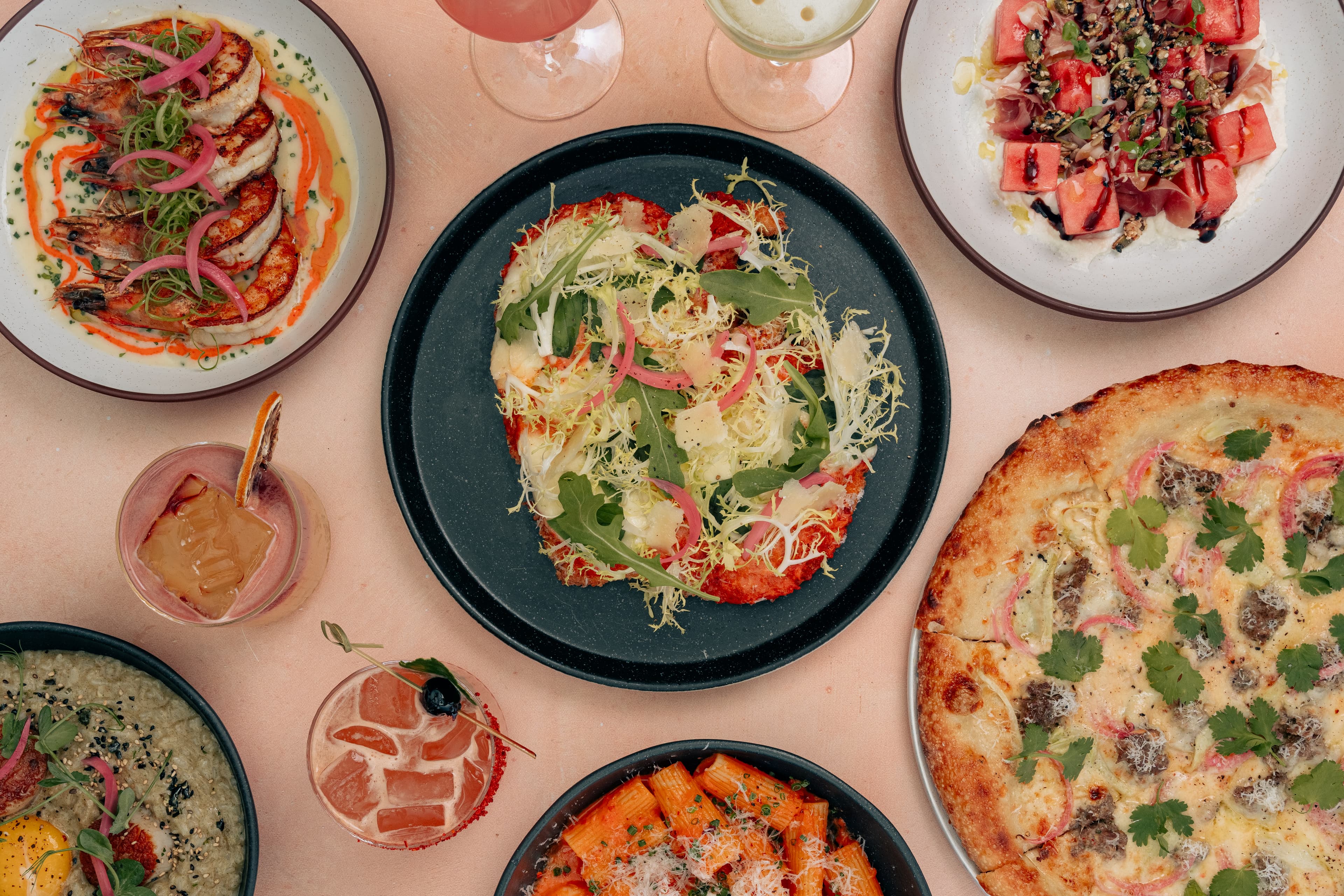 An overhead spread from INI Ristorante featuring chicken katsu parmesan, shrimp rigatoni alla rosa, mala lamb pizza topped with red onions and cilantro, watermelon and prosciutto salad, grilled shrimp dishes, cocktails, and other modern Italian-inspired plates.
