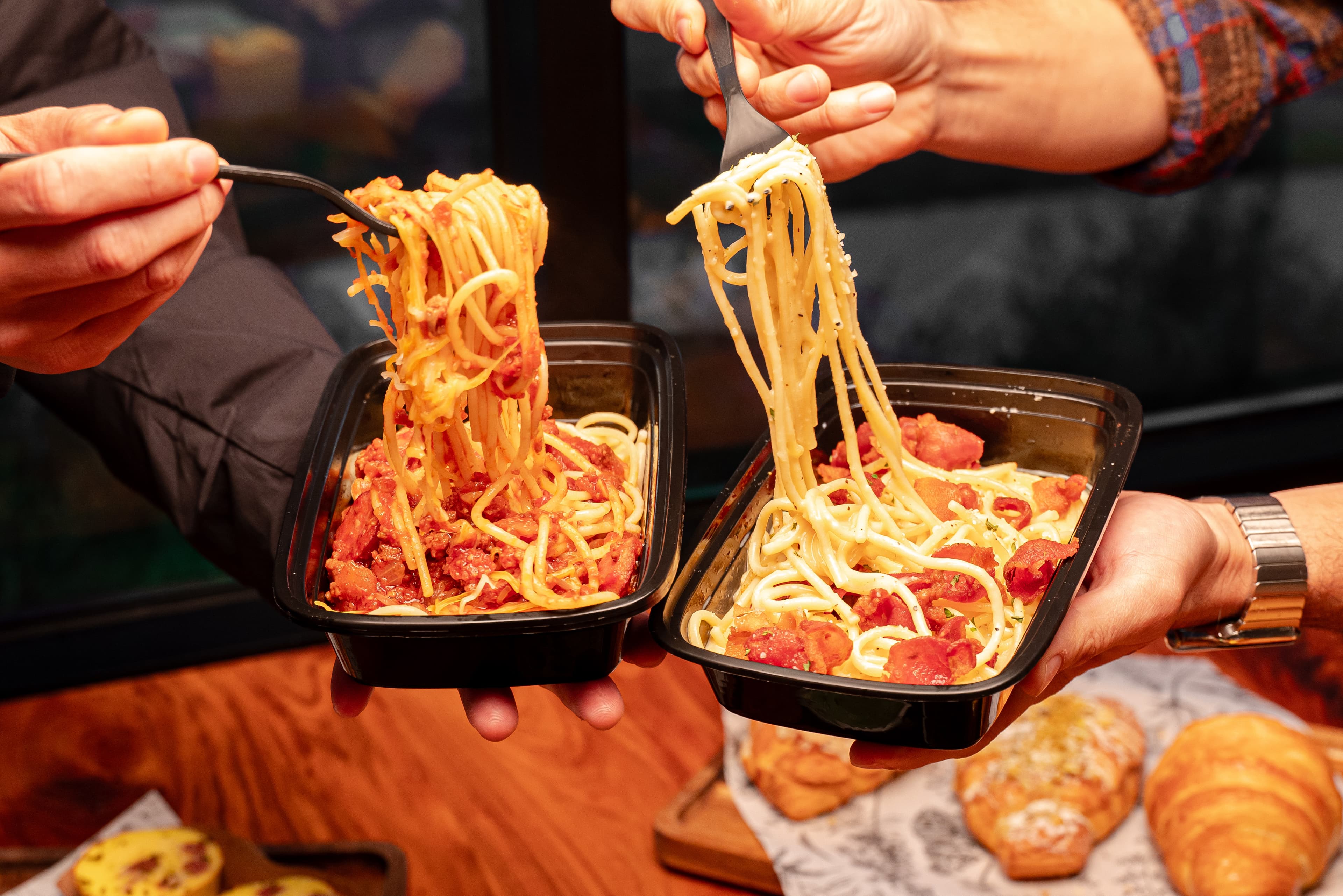 Two people holding takeout pasta dishes from INI Ristorante, featuring creamy bacon carbonara and sweet bolognese spaghetti, with forks lifting the noodles above the containers.