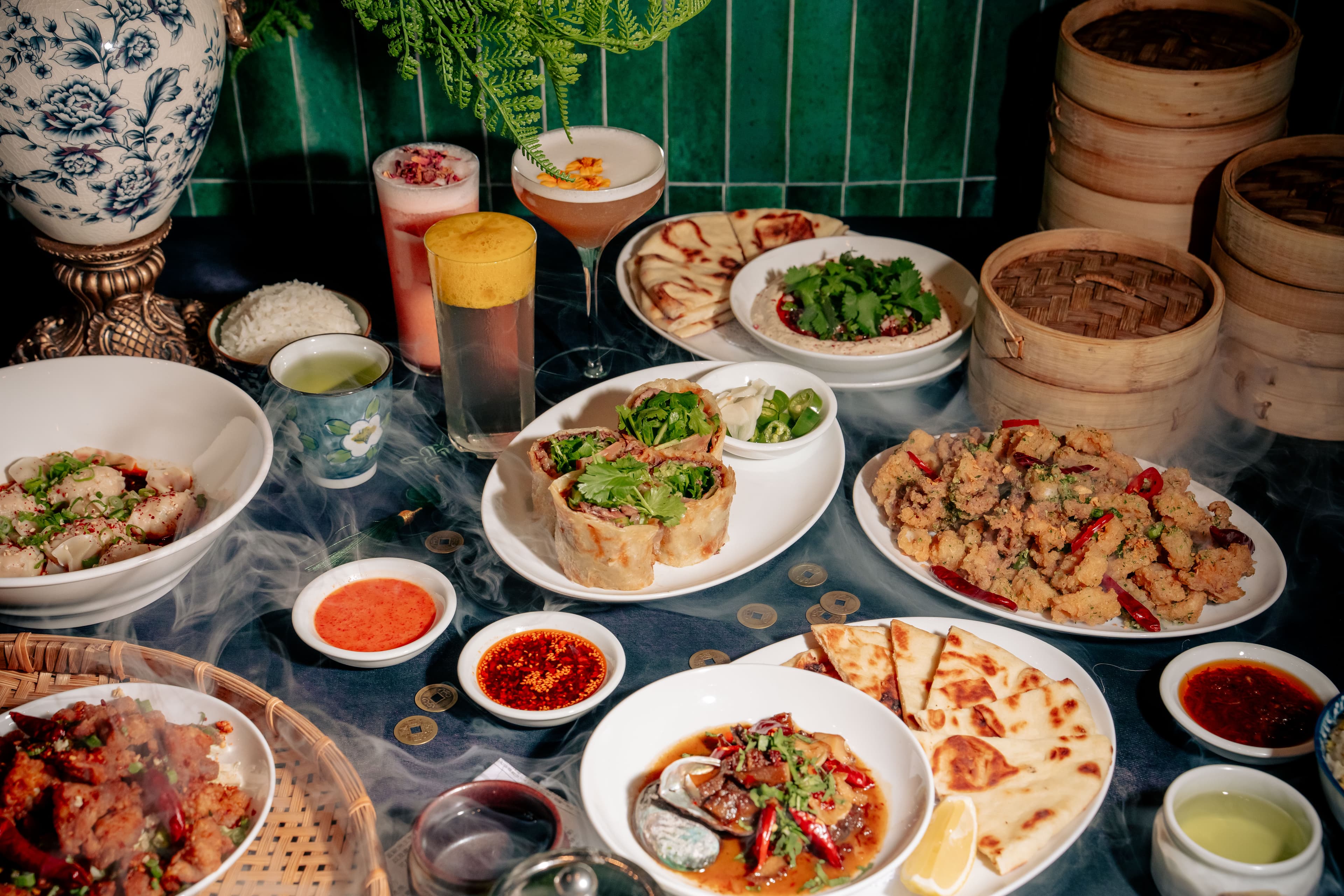 A dramatic spread from QUA Contemporary Chinese Cuisine featuring beef roll, mapo lamb dip with naan bread, calamari, abalone, spicy wontons, and cocktails surrounded by smoky presentation effects and bamboo steamers.