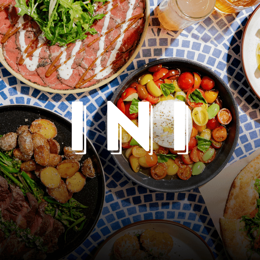INI Ristorante logo overlaid an overhead spread from INI Ristorante featuring burrata caprese with heirloom tomatoes and basil, miso picanha steak with roasted potatoes and broccolini, beef carpaccio topped with arugula and sauce drizzle, pizza, and assorted Italian-inspired dishes arranged across a blue mosaic table.